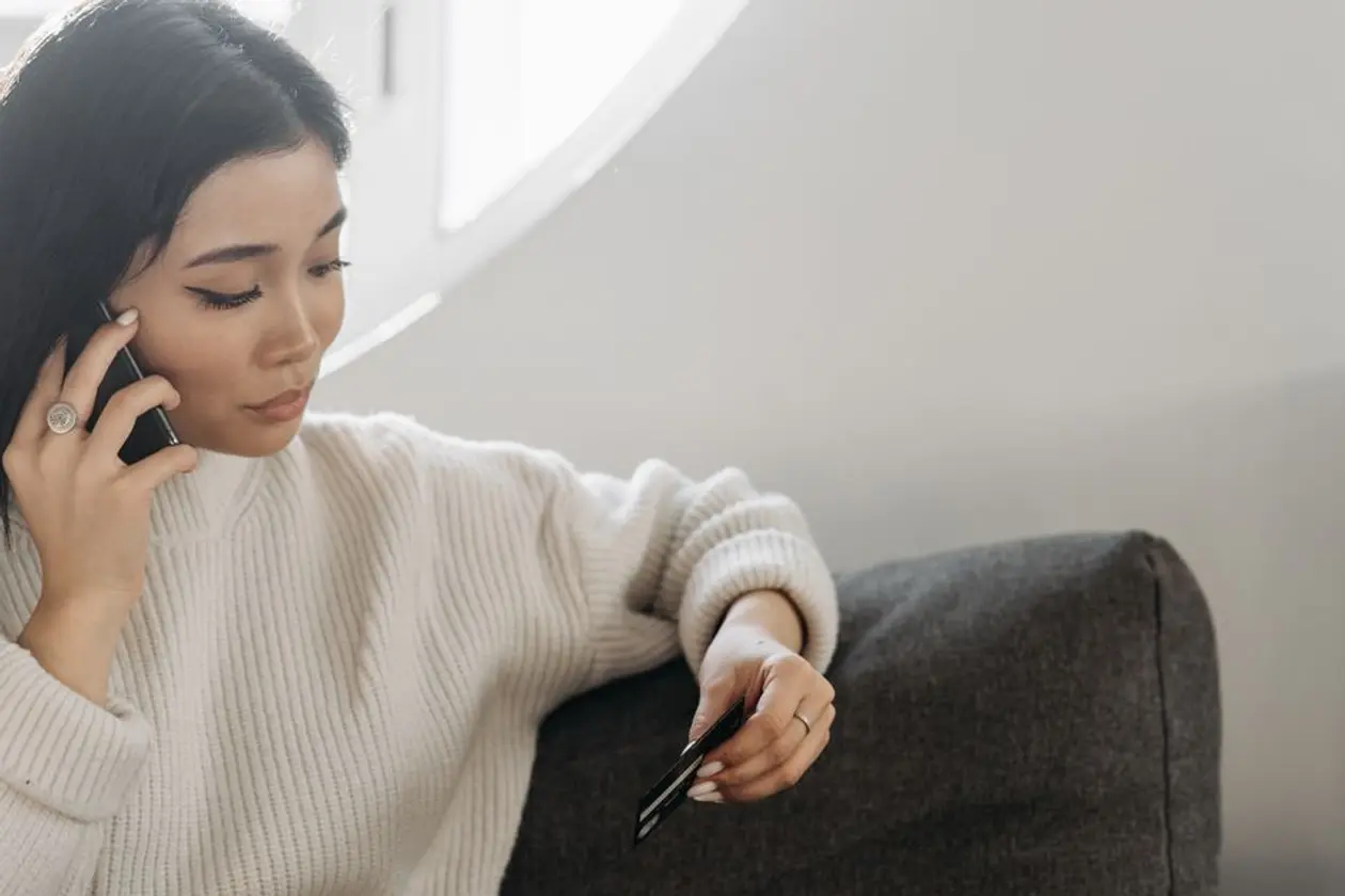 穿白色針織毛衣的亞洲女性坐在灰色沙發上講電話，另一手拿著一張卡片，示意比較信用卡分期和無卡分期前先向業者問清楚費用與條款
