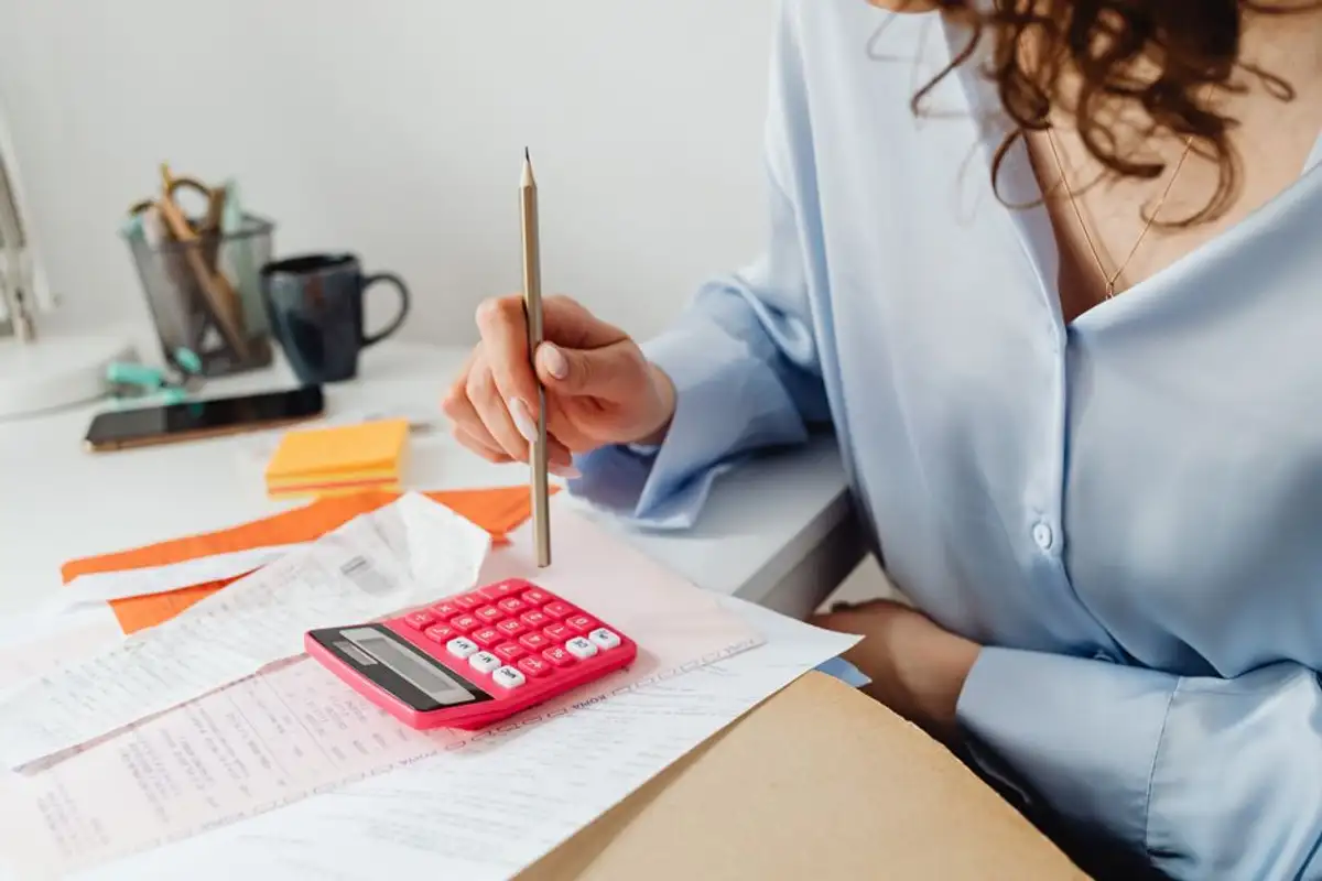 女性坐在書桌前一手握著鉛筆、一手按著粉紅色計算機，桌上散放收據與帳單，示意逐項核對本金、利息與費用後再決定清償方式