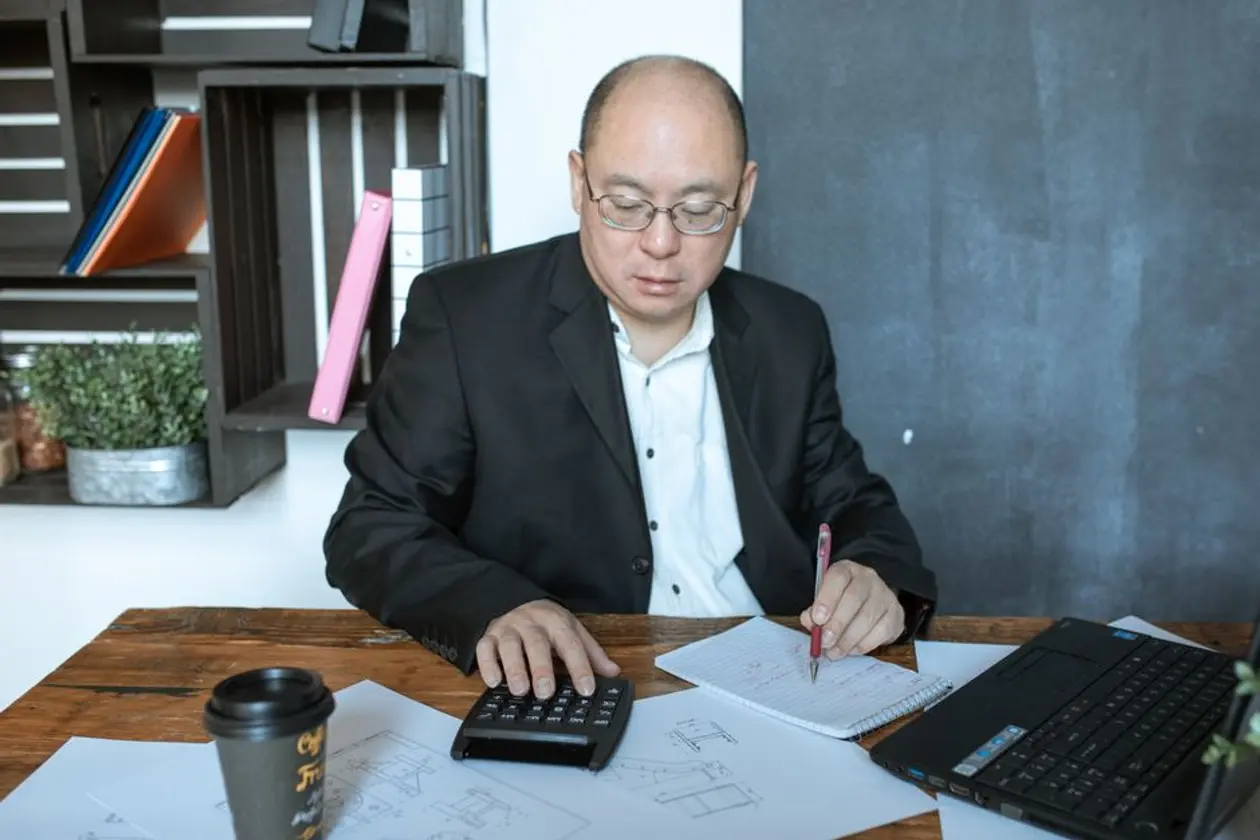 戴眼鏡的亞洲男性坐在木桌前，一手按計算機、一手在筆記本上記錄數字，桌上攤著平面圖與筆電，示意試算房貸利率與違約金