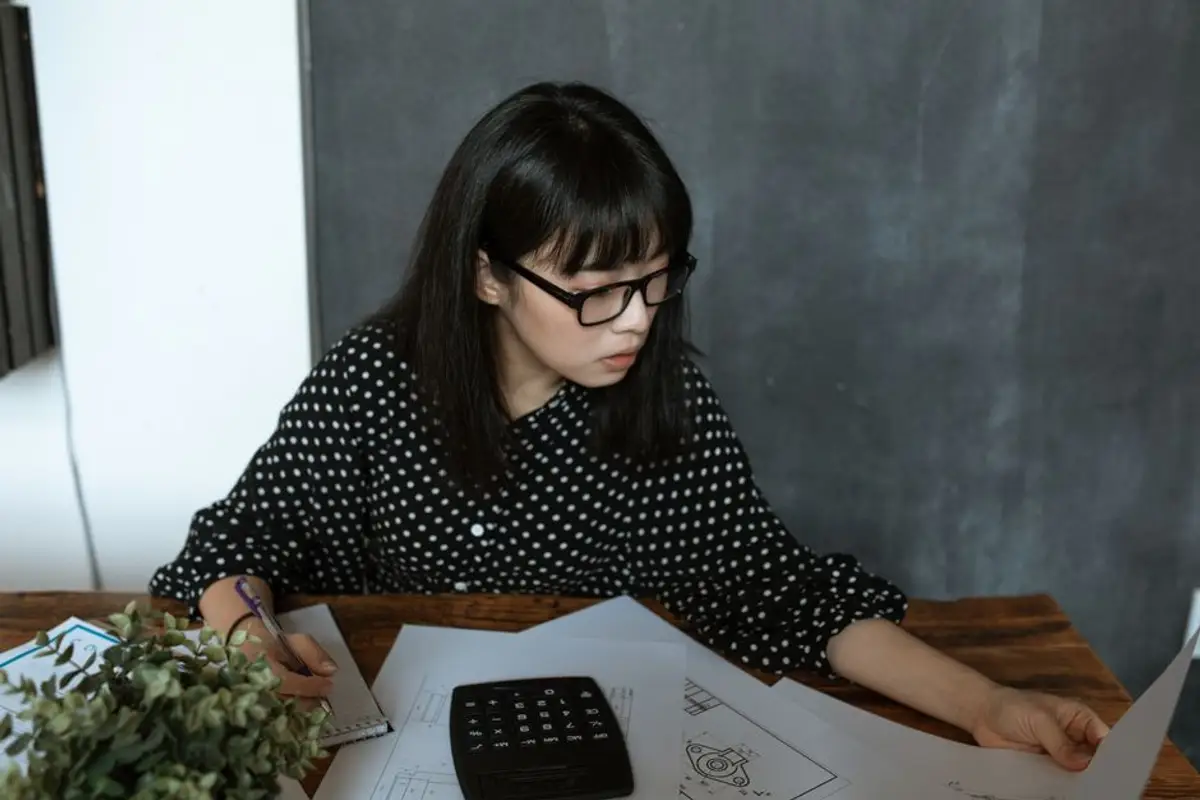 戴黑框眼鏡、穿圓點上衣的女性坐在木桌前，一手拿筆一手翻看攤開的文件，桌上放著計算機，像在逐項核算租約與還款金額