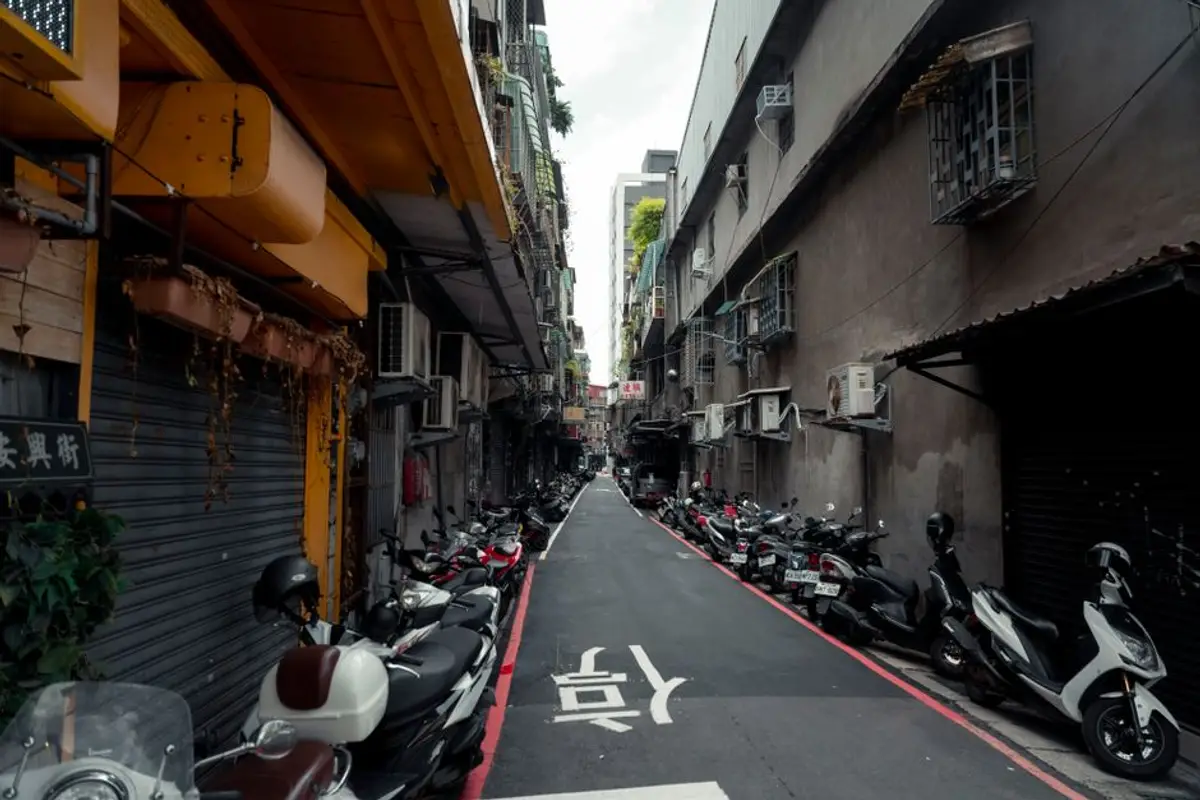 城市巷弄兩側沿著紅線停滿一整排機車，路面畫著白色轉向箭頭，兩旁是拉下鐵捲門的店面與老公寓，呈現機車通勤族日常停車的景象