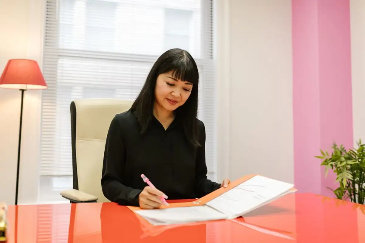 留著黑色長髮與瀏海的亞洲女性坐在明亮辦公室的紅色桌前,手拿粉紅色筆在文件夾內的書面資料上填寫,像在準備拋棄繼承的書狀與附件