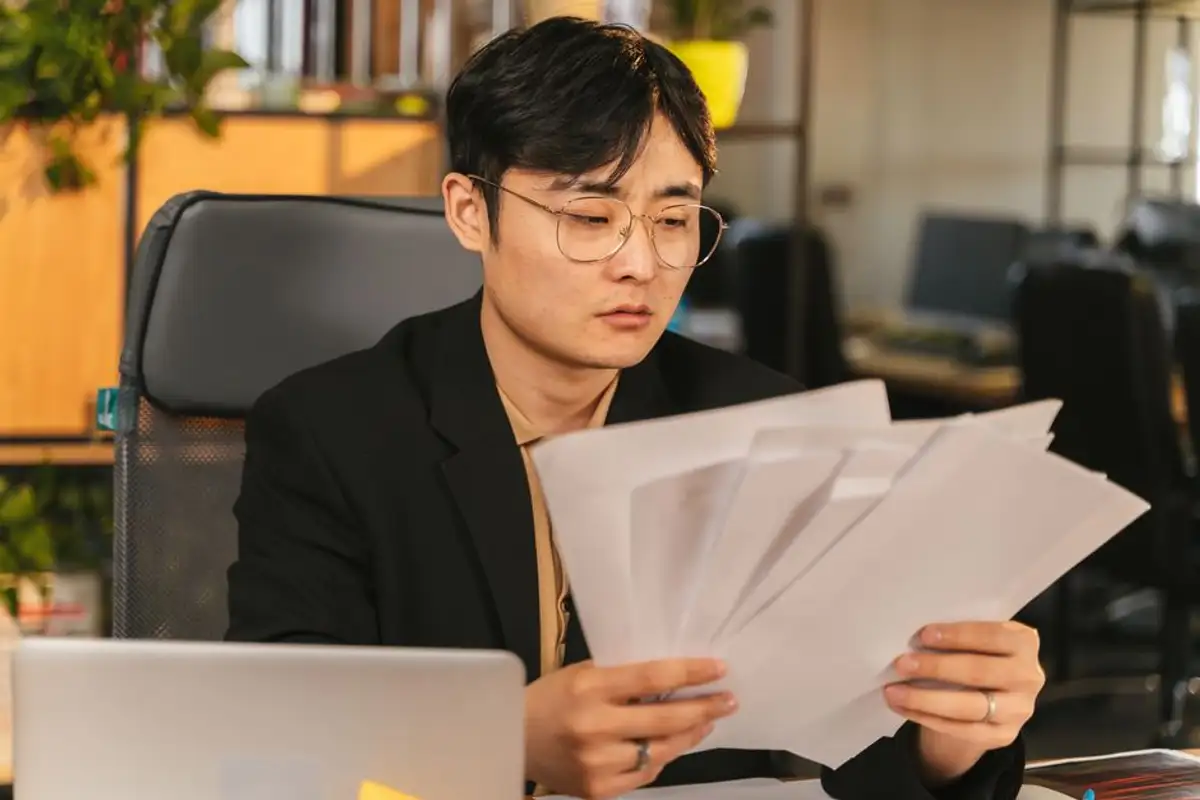 戴金屬框眼鏡、穿黑色西裝外套的亞洲年輕男子坐在辦公桌前,雙手攤開一疊文件逐張核對,像在整理父母留下的帳單與借款資料