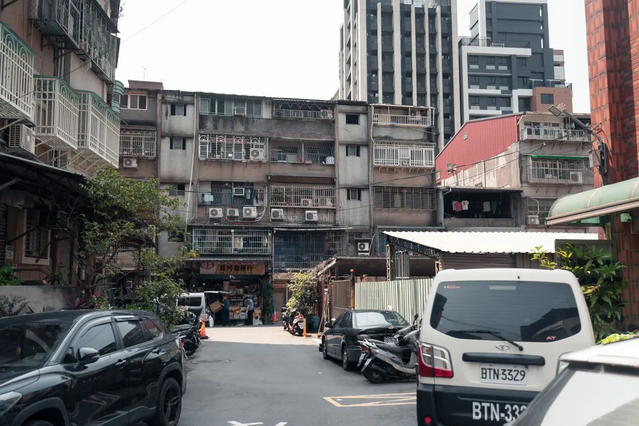 台北巷弄裡一排老舊公寓，陽台裝著鐵窗和冷氣機，一樓是店家，巷內停著汽車和機車，後方是新建大樓，示意父母名下的公寓也可能成為借款擔保品