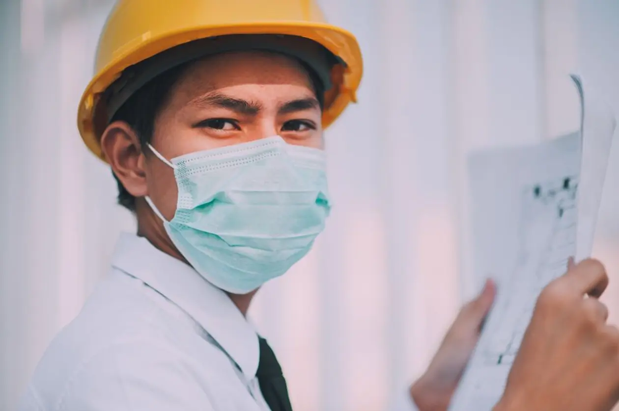 戴黃色安全帽和口罩的亞洲年輕工程師一手拿著建築圖面看向鏡頭，示意自地自建要先取得建造執照並依工程進度分段撥款
