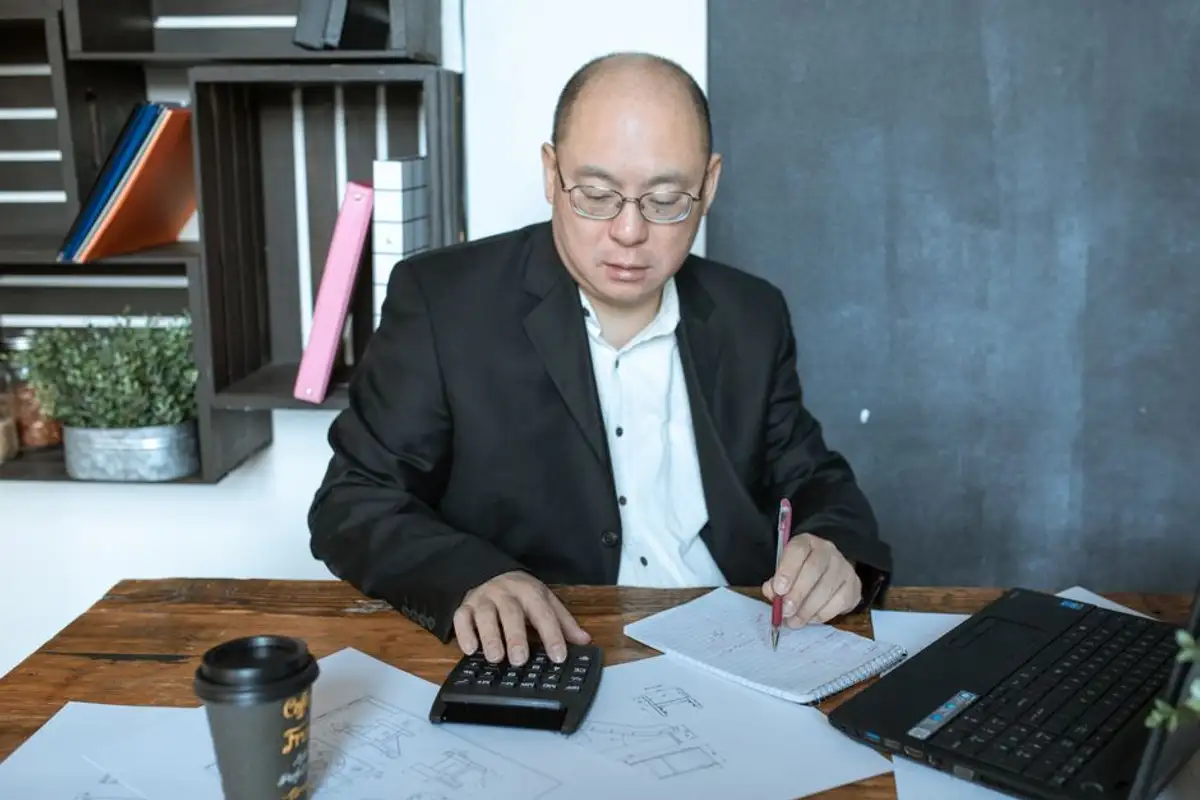 戴眼鏡的亞洲中年男子坐在木桌前，一手按計算機、一手拿紅筆在筆記本上記錄，桌上有筆電、外帶咖啡杯與文件，像在試算車價與剩餘車貸的差額