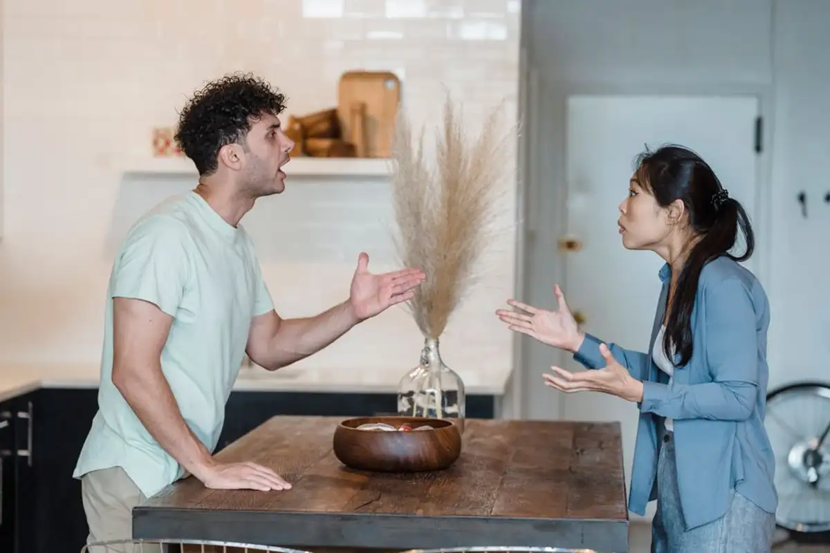 一對男女隔著廚房木桌激烈爭執,男子張口攤手、綁馬尾的亞洲女子也伸手回應,桌上放著木碗與乾燥花,呈現夫妻為債務與財產歸屬起爭執的情景