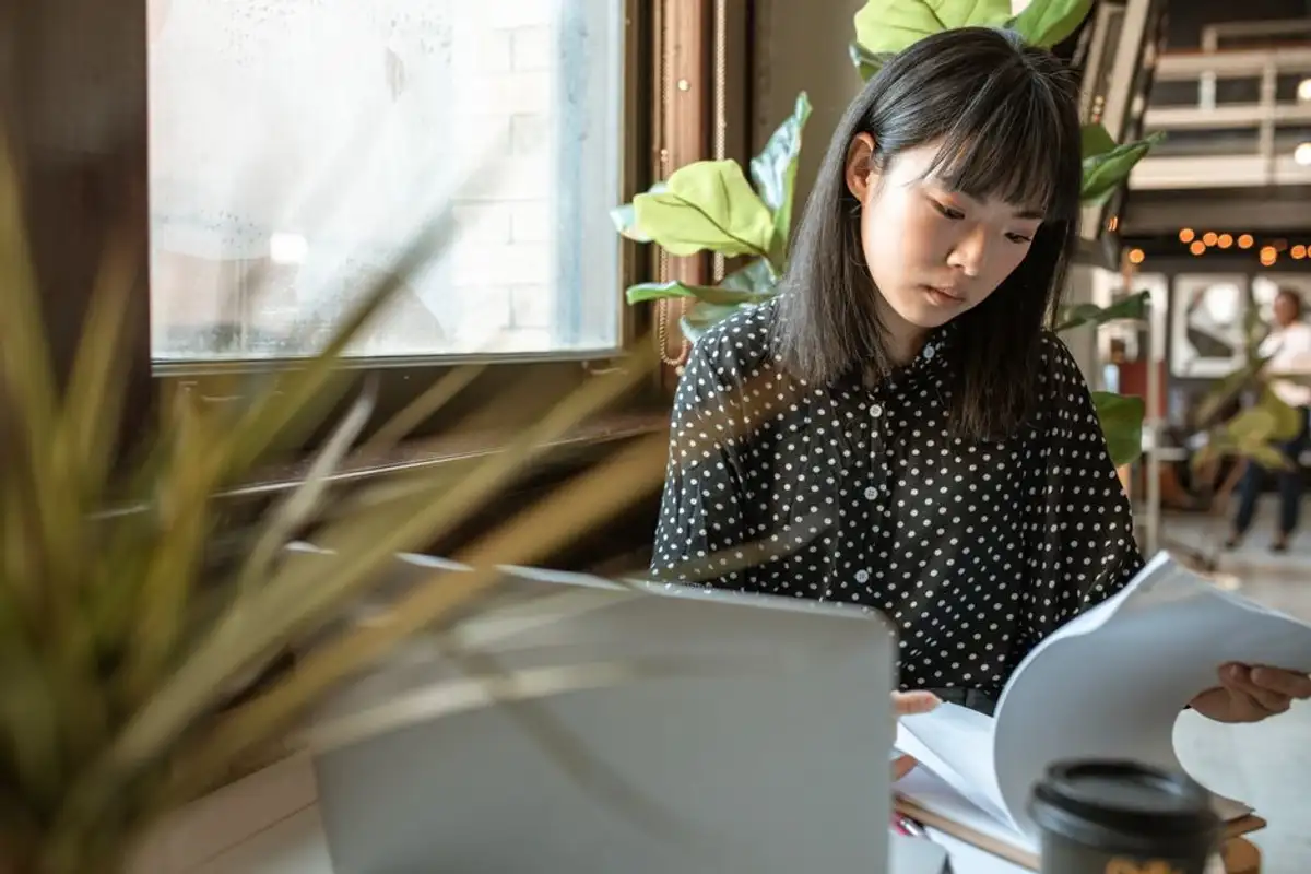 一位留著齊瀏海短髮的亞洲女性坐在窗邊桌前,低頭翻閱一疊文件,桌上有筆電與外帶咖啡杯,像在整理自己名下財產的所有權證明
