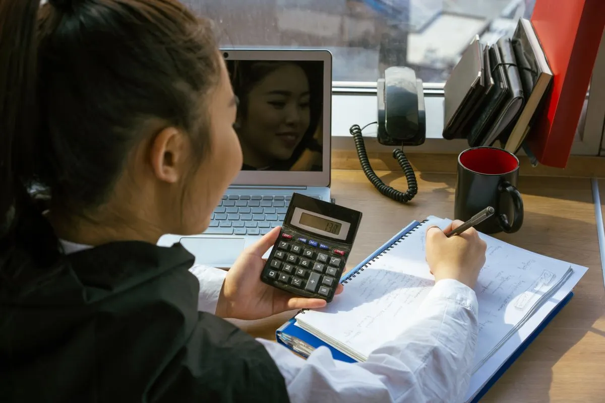 綁馬尾的女性坐在窗邊辦公桌前,一手拿計算機、一手在筆記本上寫字,旁邊有筆電與電話,像在試算不同期數的信貸月付金