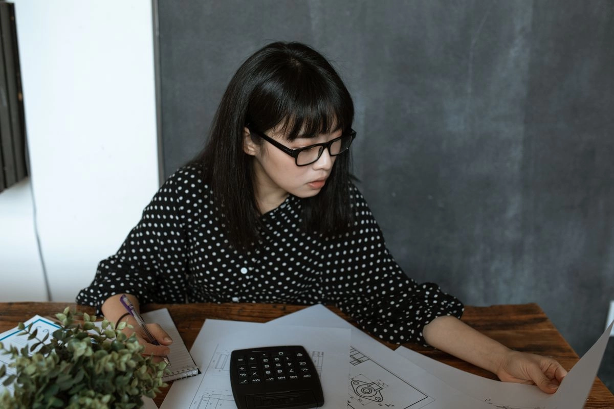 戴黑框眼鏡的亞洲女性坐在桌前翻閱文件並寫筆記，桌上放著計算機與盆栽，像在整理申請信貸要準備的財力資料