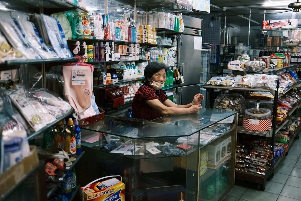 戴口罩的年長女性站在台灣傳統雜貨店的玻璃櫃台後方，貨架上擺滿零食與日用品，對應店舖負責人貸款的申請族群