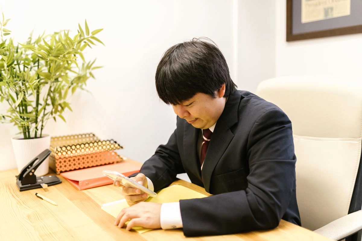 穿西裝的亞洲男性坐在辦公桌前滑手機,桌上放著黃色文件夾,準備用手機上傳財力證明申請渣打信貸