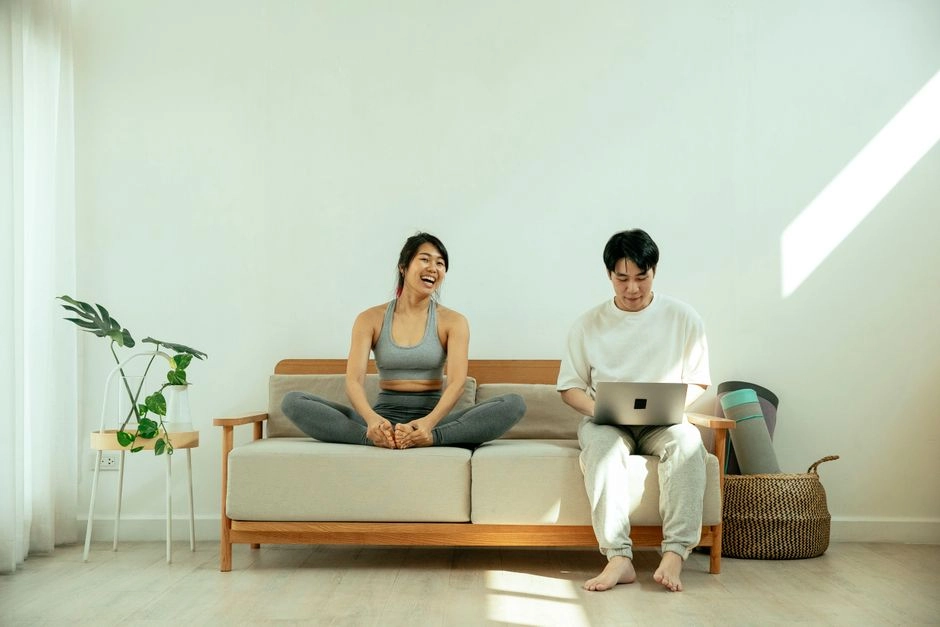 年輕亞洲夫妻坐在客廳木框沙發上,先生用筆電查資料,太太盤腿坐在一旁開心大笑,呈現夫妻一起在家規劃的場景