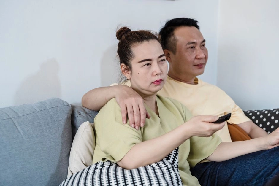 一對亞洲中年夫妻穿著淺色家居服坐在客廳灰色沙發上,先生摟著太太的肩膀,太太拿著遙控器看電視,呈現中年家庭在家的日常