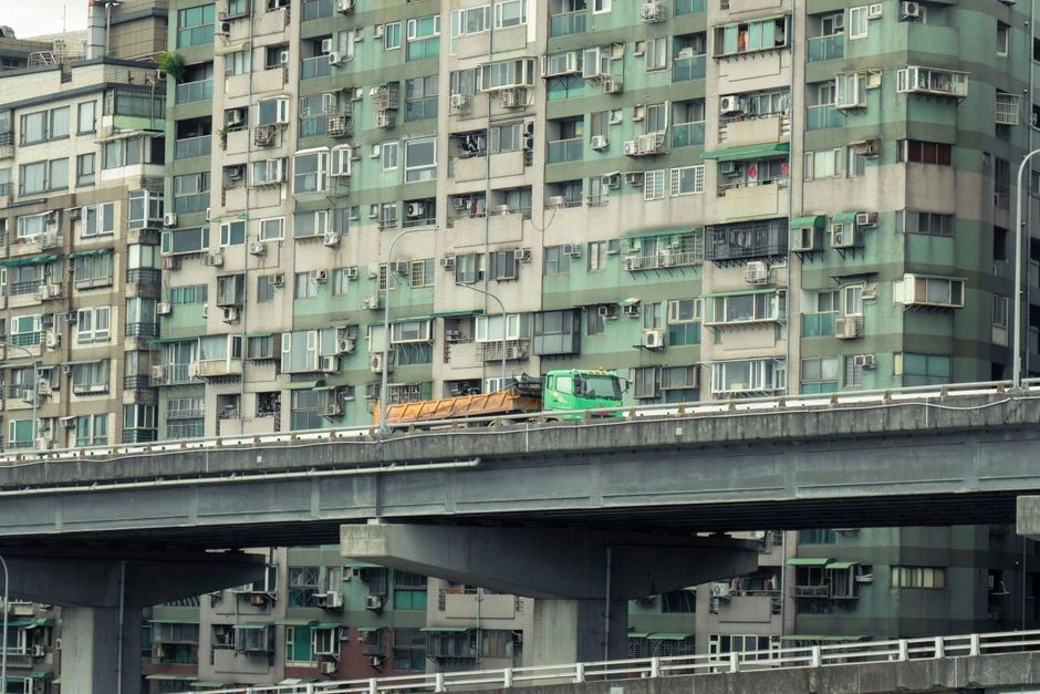 台北一排密集的公寓大樓前方,一輛綠色卡車駛過高架道路,畫面可見整面窗戶與冷氣機,呈現都市常見的住宅景象