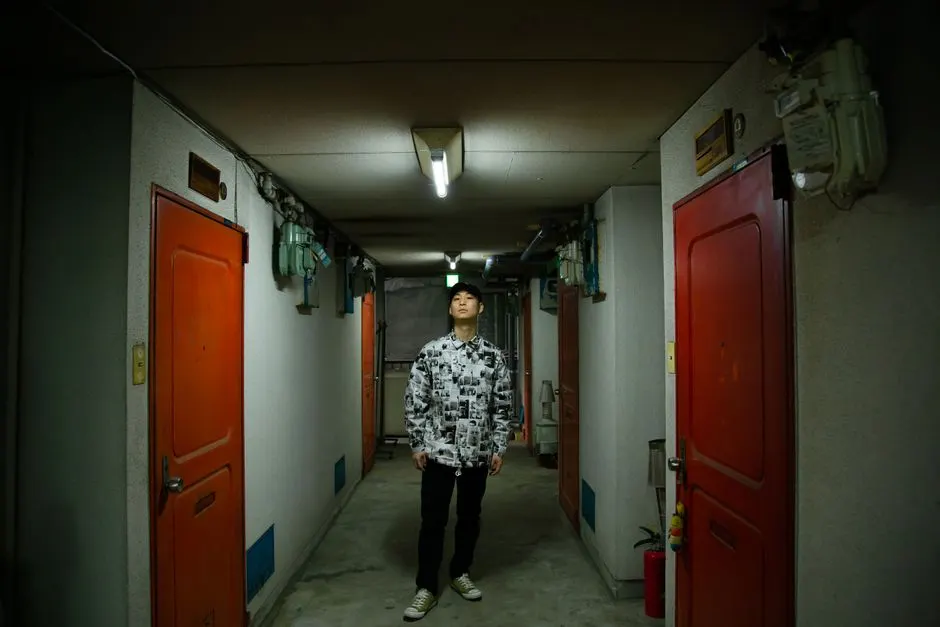 一名穿黑白圖案襯衫的年輕男子站在公寓走廊中間抬頭張望，兩側是紅色鐵門，天花板有日光燈與管線，牆上掛著電表