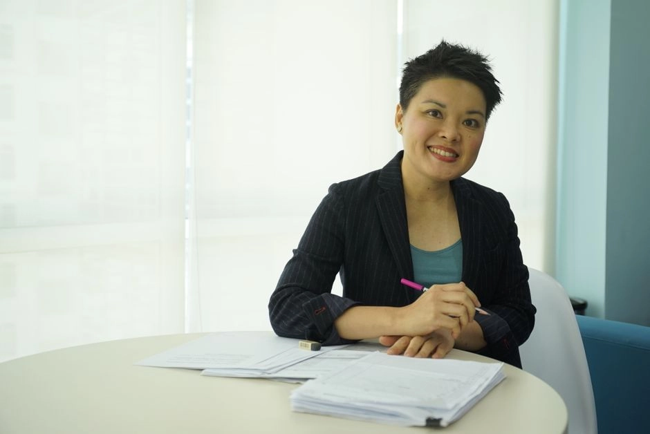 短髮女性穿西裝外套坐在圓桌前，手握筆、面前放著一疊文件微笑