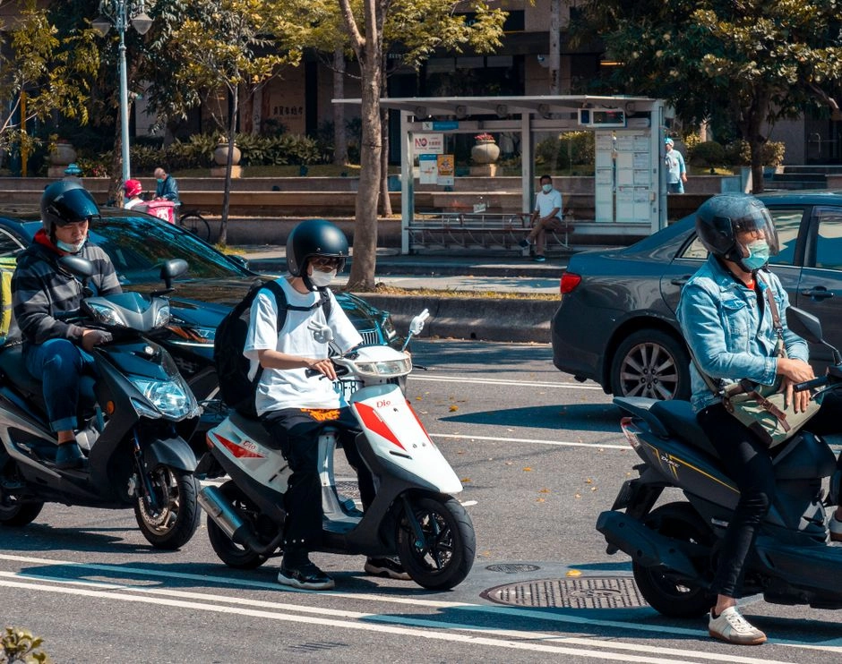 戴口罩和安全帽的機車騎士們在市區路口停等，中間一名穿白色上衣、背黑色後背包的騎士騎著紅白相間的機車，旁邊有轎車與公車站